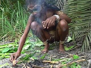 African native woman cooks topless in wild jungle setting. Her skinny frame shines under the sun.